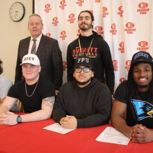 All four students will play football at the Division 2 and Division 3 level, going to college on scholarships. Pictured behind them are Superintendent of Schools William Hart and Head Coach Justin Flores.