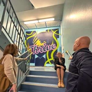 Malden Public Schools’ third-year Superintendent, Dr. Ligia Noriega-Murphy (seated), admires the newest artistic creation to the Malden High School walls, a mural that is near completion on the stairwell between the first and second floors of Boyle House. The mural expresses the present school theme, “This is Our Year to Thrive,” and features the word theme surrounded by the school mascot, “Nedlam,” the lion. The wall mural was designed and painted by Malden Public Schools art students supervised and assisted by Malden High art teacher Joseph Luongo. Shown above, also admiring the art creation, are Malden High School Principal Chris Mastrangelo (at right) and Assistant Superintendent Emilys Peña (at left). (Advocate Photo)