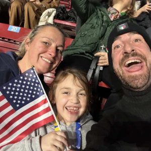 ROOTING ON THE USA: Seven-year-old Pia Mercurio waved a miniature American flag, and she and her parents – Jackie and Scott Mercurio – cheered at an event in Milano, Italy, where they watched the 2026 Winter Olympic Games. (Courtesy photo to The Saugus Advocate)